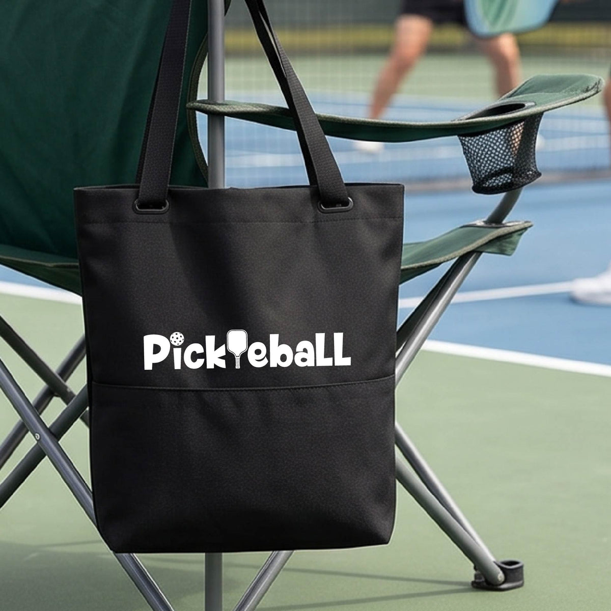 A close-up of a black medium size tote bag with a white graphic text 'PICKLEBALL' on the leg with a pickleball paddle replacing the L, on a pickleball court.