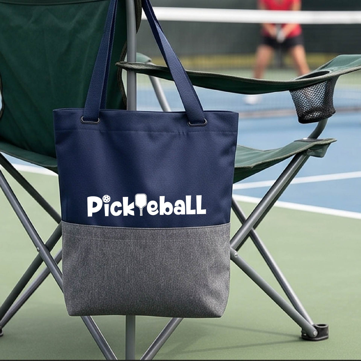 A close-up of a navy blue and gray medium size tote bag with a white graphic text 'PICKLEBALL' on the leg with a pickleball paddle replacing the L, on a pickleball court.