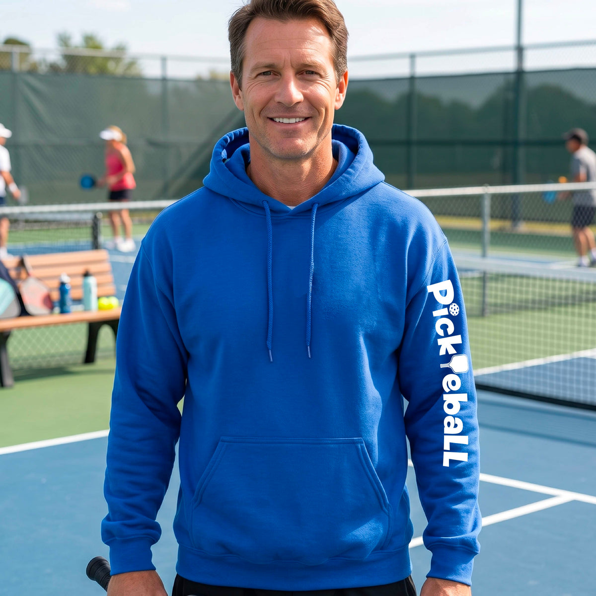 A smiling man wearing a bright blue pullover hoodie with 'Pickleball' printed vertically in white on the left sleeve, standing on a blue pickleball court with other players and a fence in the background.