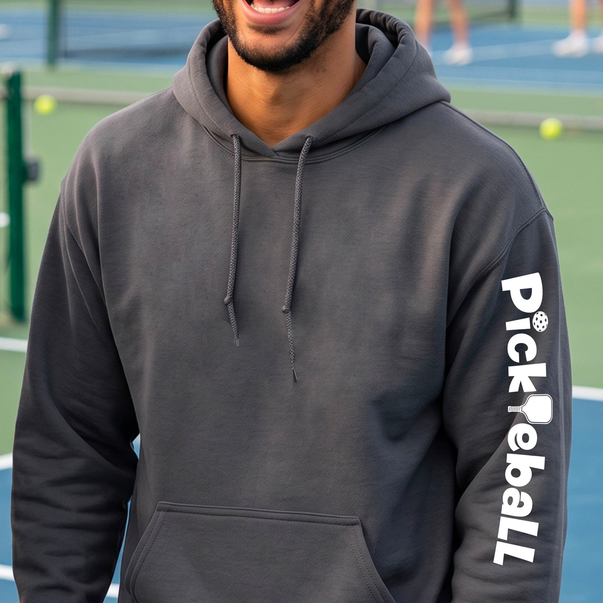 A man on a pickleball court wearing an gray hoodie, shown from the back. The hoodie features the word 'Pickleball' printed vertically in white, with a pickleball paddle graphic replacing the letter 'i'.