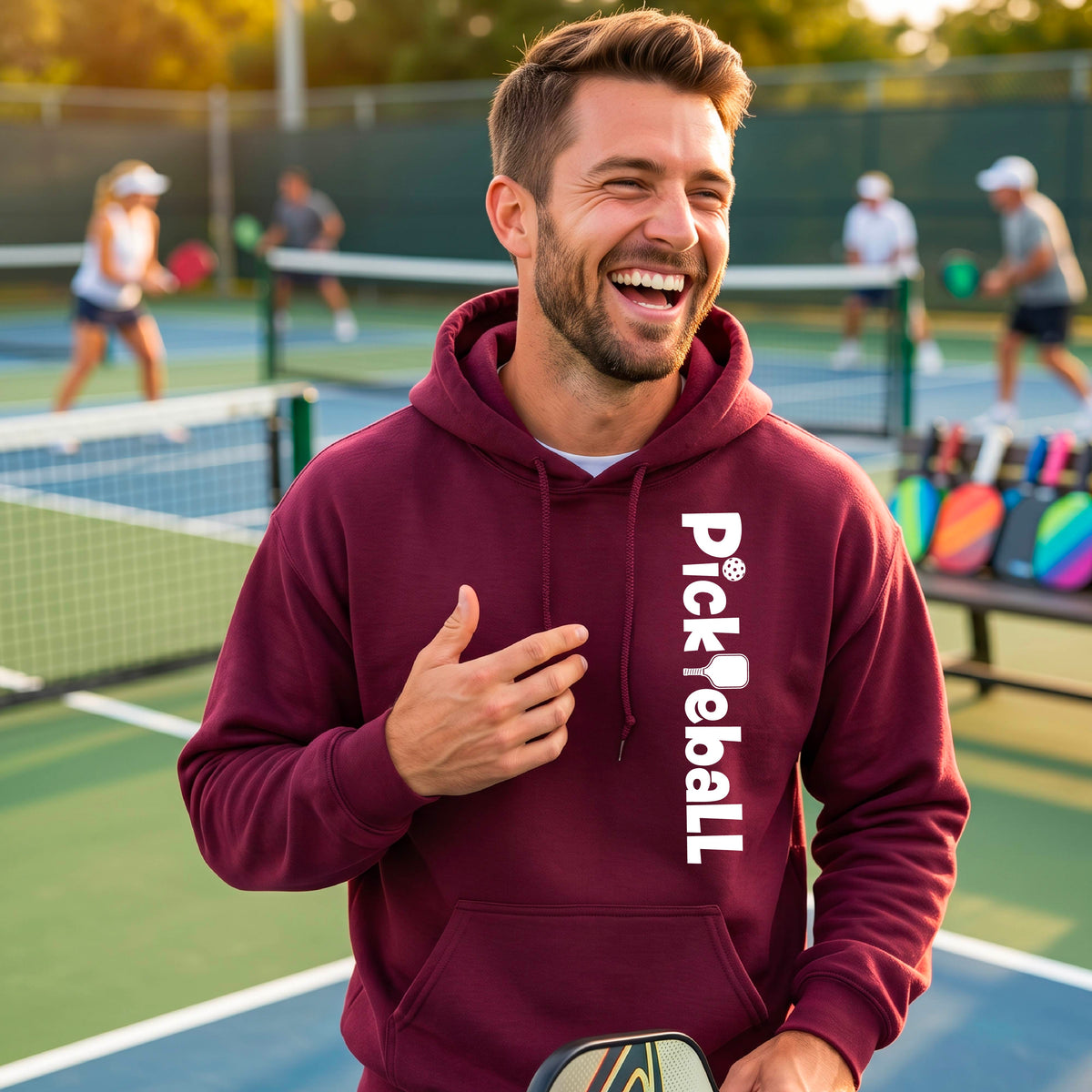 A man on an outdoor pickleball court wearing a maroon hoodie with 'PickReball' printed in white vertically on the chest, holding a pickleball paddle. Other players are visible in the background.