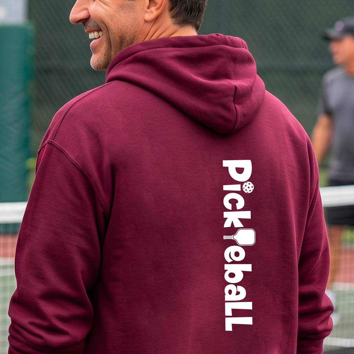 A man on a tennis court wearing a maroon hoodie with 'Pickleball' printed vertically in white on the back.