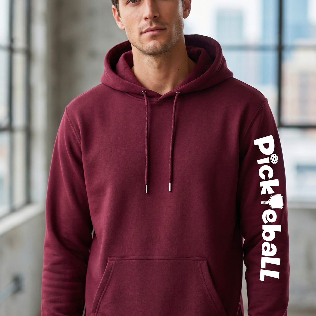 A man wearing a maroon pullover hoodie with a white vertical 'Pickleball' graphic on the right sleeve, standing in a bright indoor setting with blurred windows in the background.