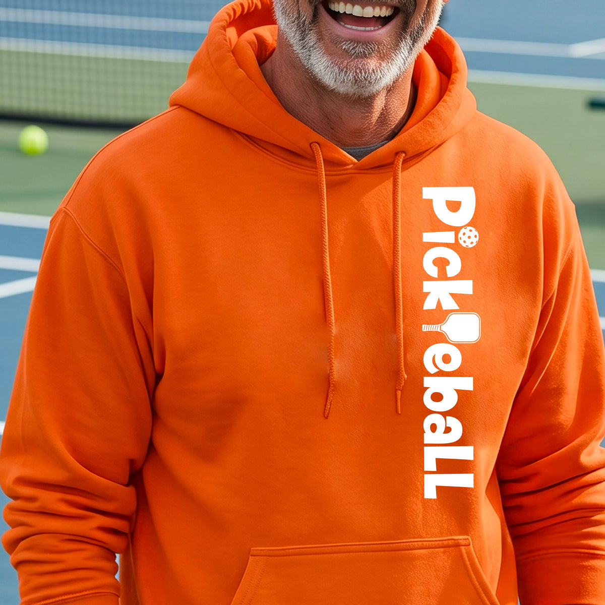A man wearing a bright orange hoodie with a vertical white 'Pickleball' logo printed on the right side, standing on a blue and green pickleball court.