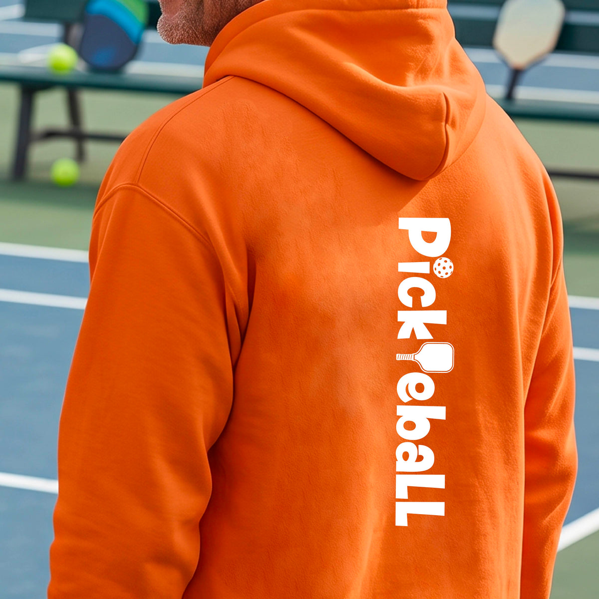A man on a pickleball court wearing an orange hoodie, shown from the back. The hoodie features the word 'Pickleball' printed vertically in white, with a pickleball paddle graphic replacing the letter 'i'.