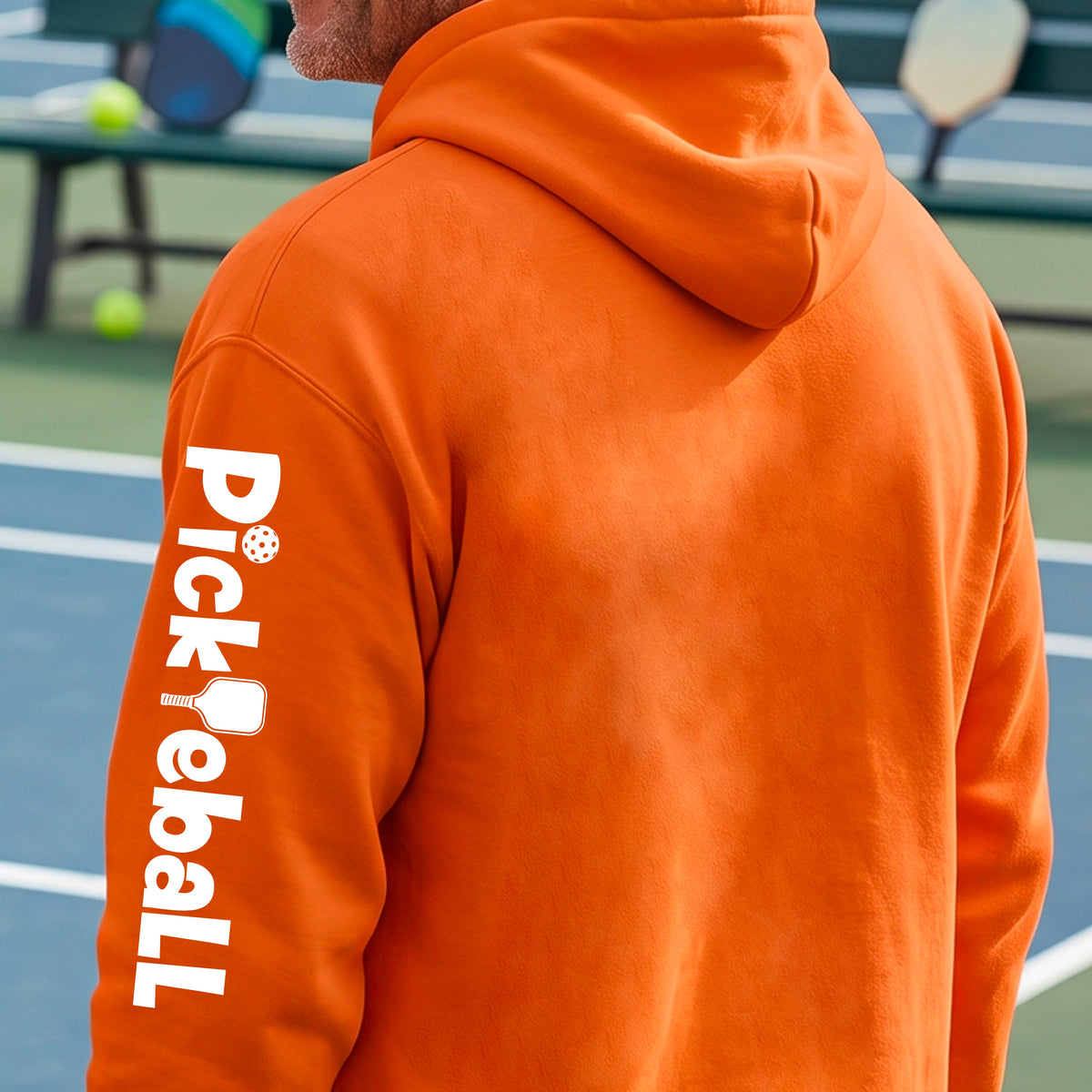 A man on a tennis court wearing an orange hoodie, shown from the back and side. The hoodie features the word 'Pickleball' printed vertically in white down the left sleeve, with a pickleball and paddle graphic.