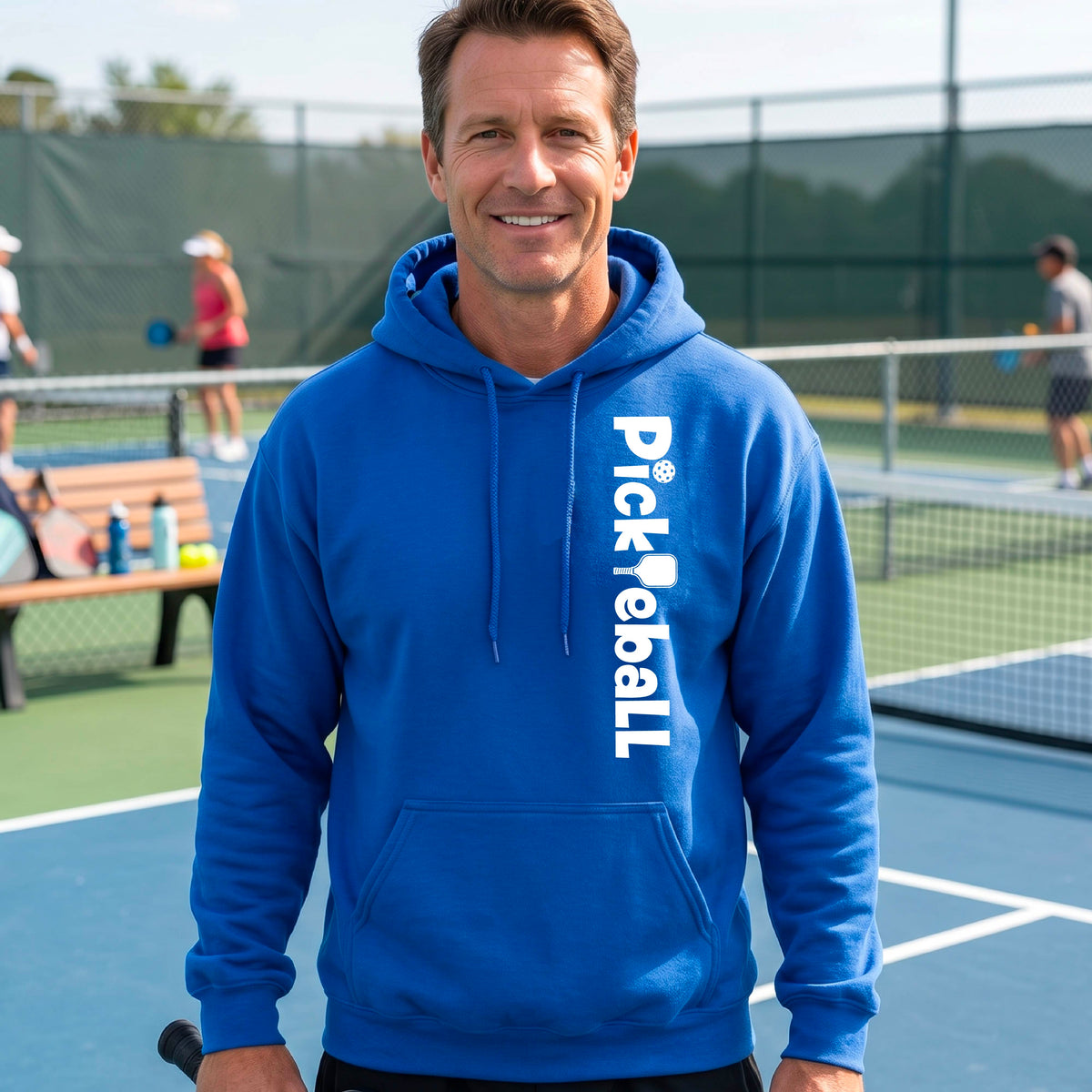 A smiling man wearing a bright blue pullover hoodie with 'Pickleball' printed vertically in white on the front, standing on an outdoor pickleball court.