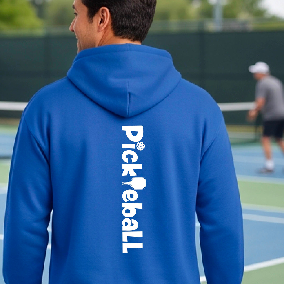 A man viewed from behind wearing a royal blue hoodie with 'Pickleball' printed vertically in white on the back, standing on a pickleball court.