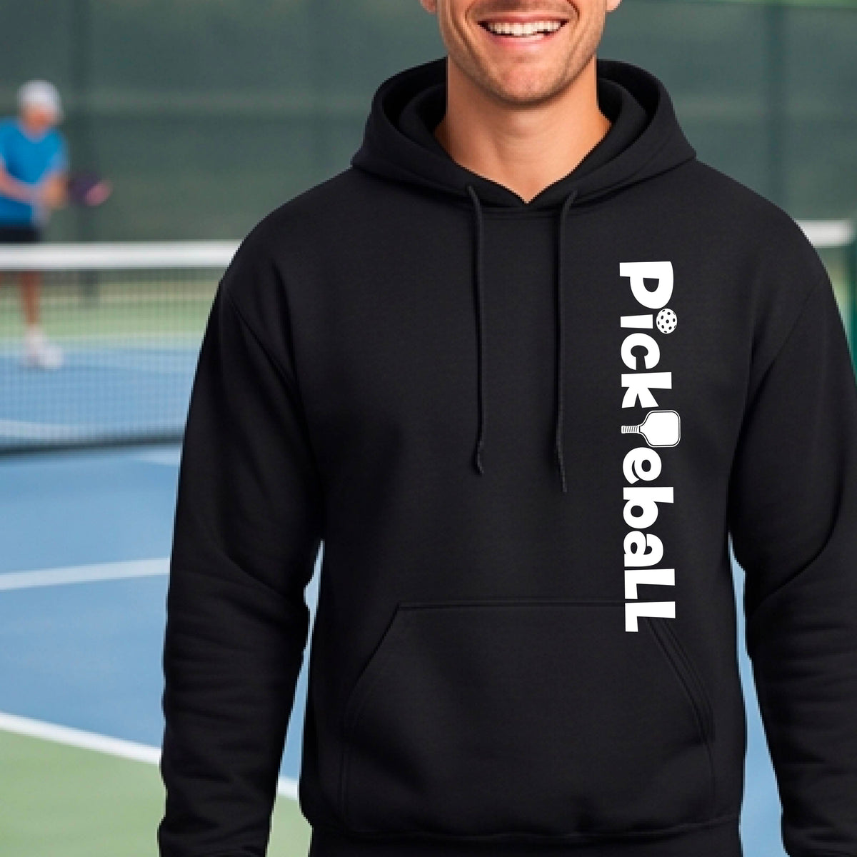 A man wearing a black pullover hoodie with 'Pickleball' printed vertically in white on the right side of the chest, standing on a blue and green pickleball court with a blurred background.
