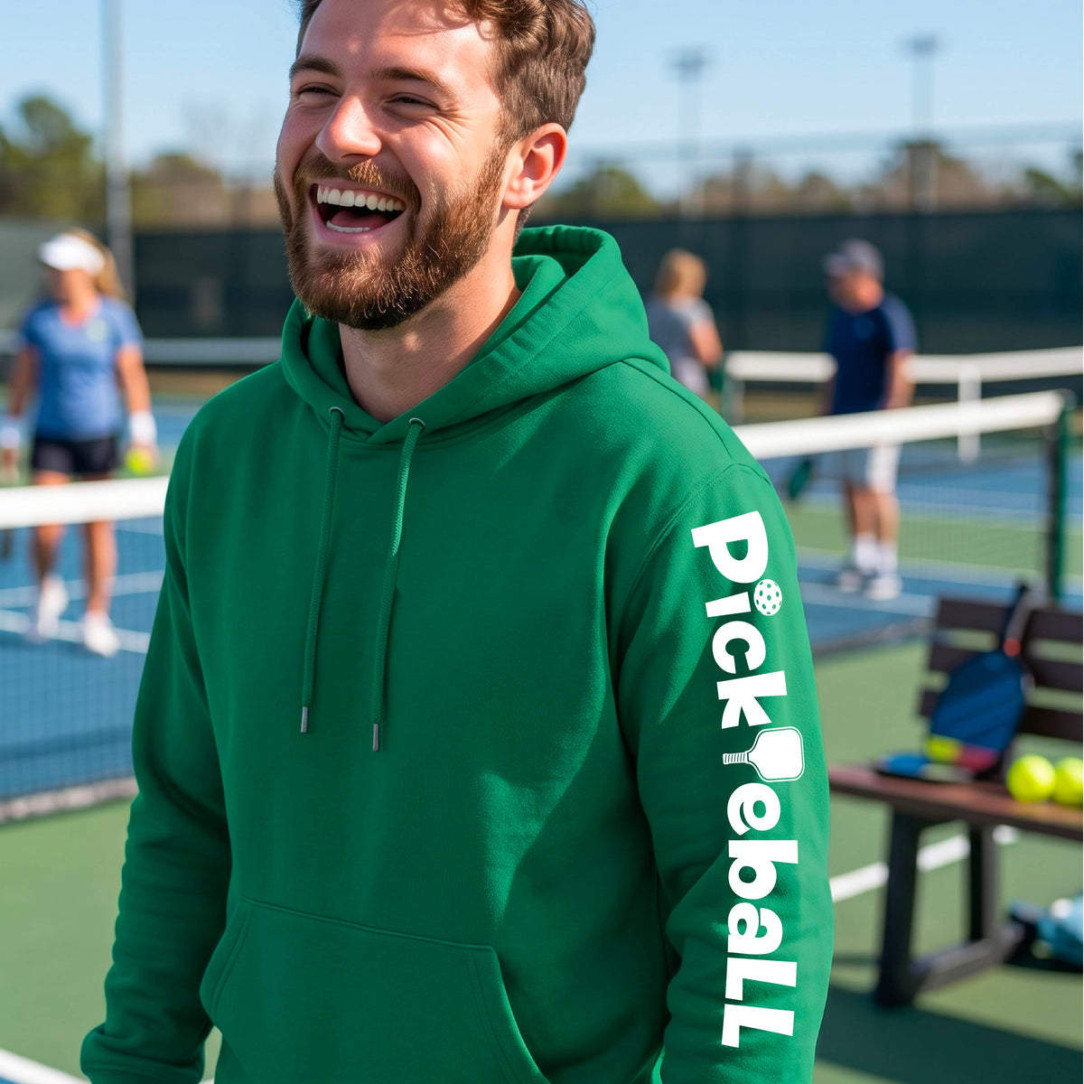 A man smiling on a pickleball court wearing a bright green hoodie with 'Pickleball' printed vertically in white on the sleeve.