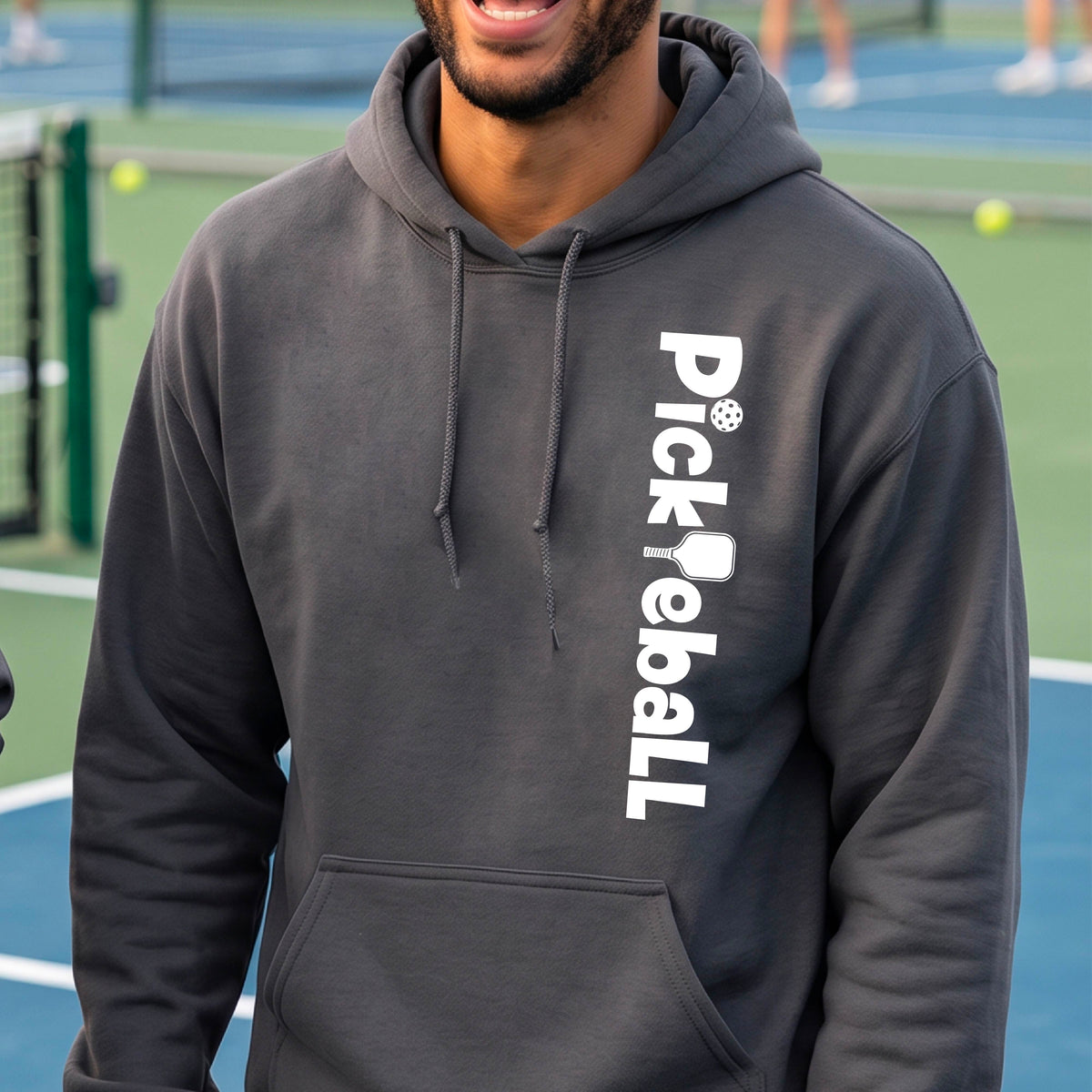 A man wearing a dark gray hoodie with 'Pickleball' printed vertically in white on the right chest, standing on a blue and green pickelball court.