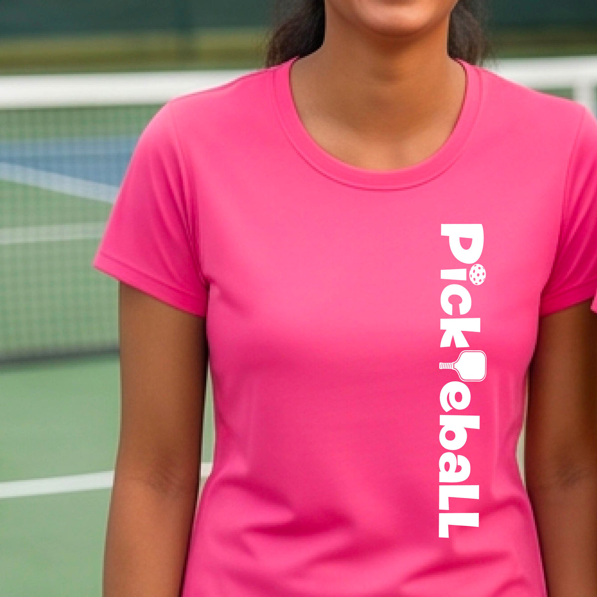 A woman wearing a pink short-sleeve t-shirt with a white graphic text 'PICKLEBALL' with a pickleball paddle replacing the L, on the front, standing on a pickleball court.