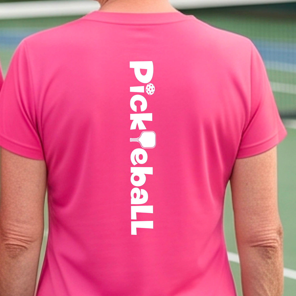 A woman wearing a pink short-sleeve t-shirt with a white graphic text 'PICKLEBALL' with a pickleball paddle replacing the L, on the back, standing on a pickleball court.