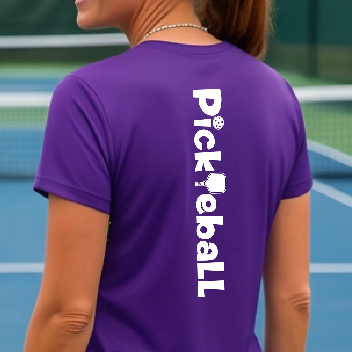 A woman wearing a purple short-sleeve t-shirt with a white graphic text 'PICKLEBALL' with a pickleball paddle replacing the L, on the back, standing on a pickleball court.