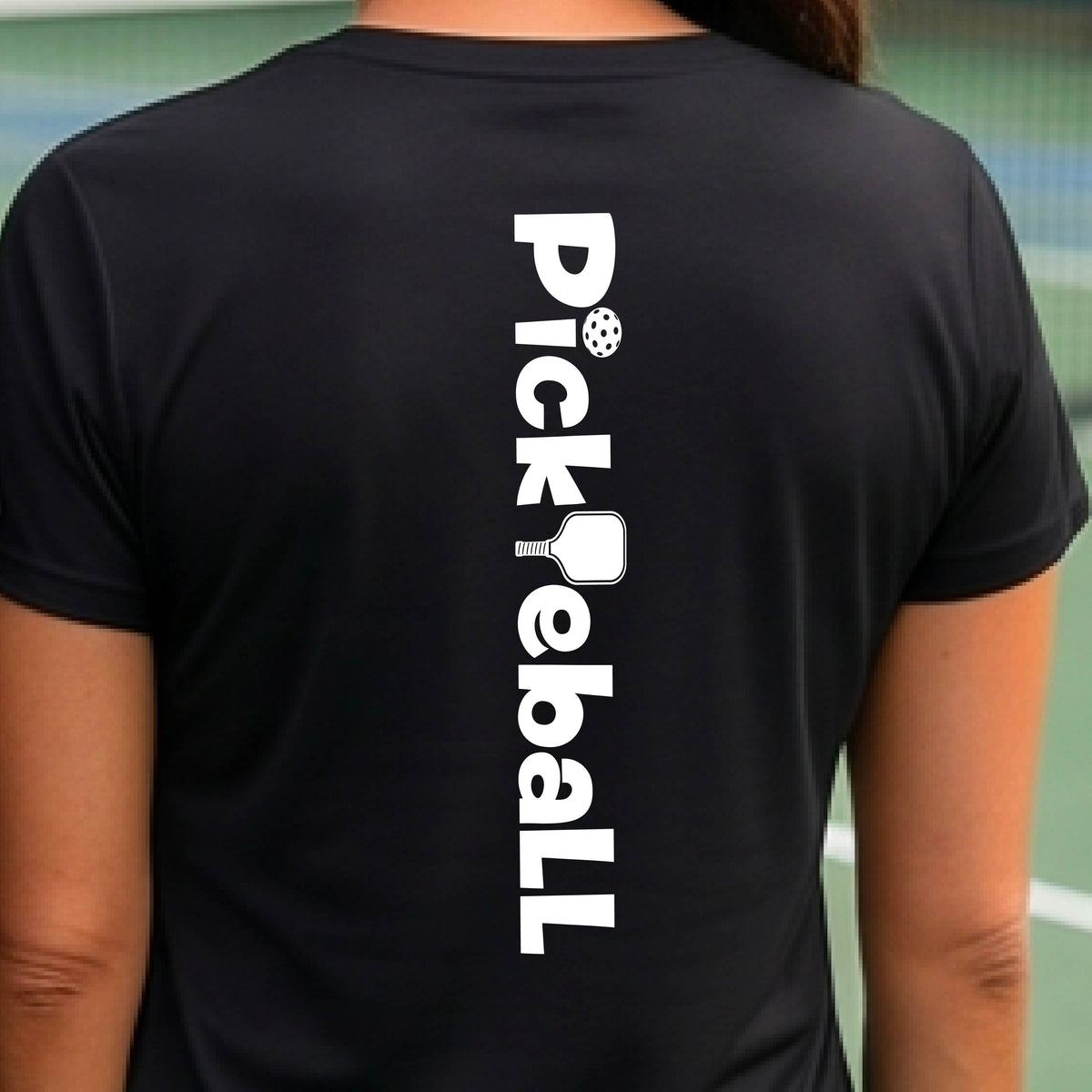 A woman wearing a black short-sleeve t-shirt with a white graphic text 'PICKLEBALL' with a pickleball paddle replacing the L, on the back, standing on a pickleball court.