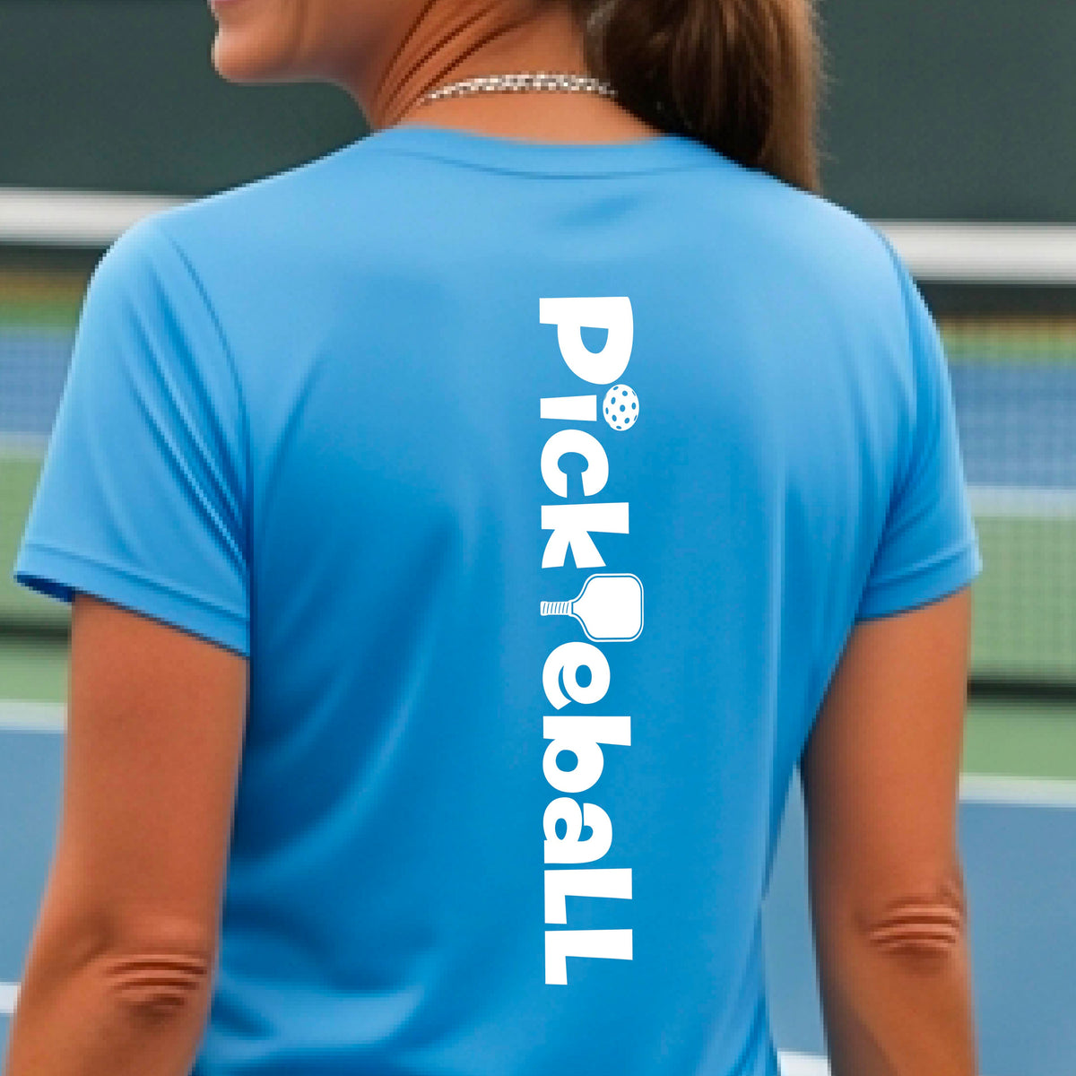 A woman wearing a blue short-sleeve t-shirt with a white graphic text 'PICKLEBALL' with a pickleball paddle replacing the L, on the back, standing on a pickleball court.