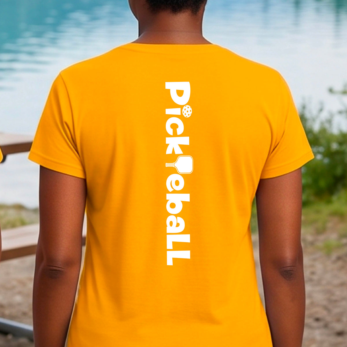 A woman wearing a gold short-sleeve t-shirt with a white graphic text 'PICKLEBALL' with a pickleball paddle replacing the L, on the back, standing on a pickleball court.