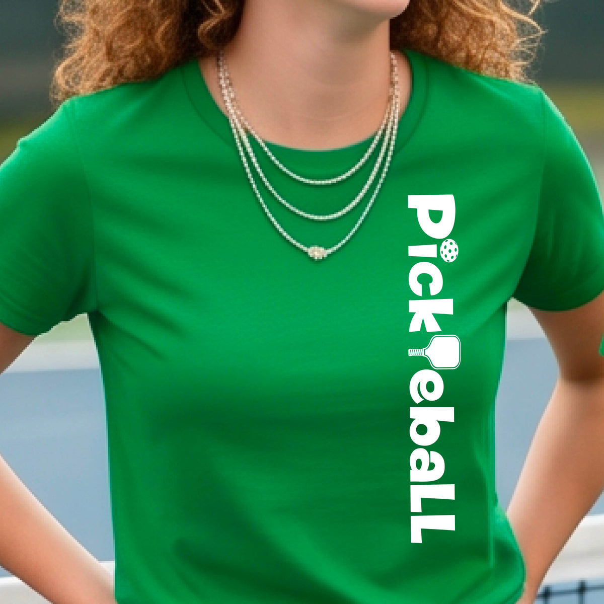 A woman wearing a green short-sleeve t-shirt with a white graphic text 'PICKLEBALL' with a pickleball paddle replacing the L, on the front, standing on a pickleball court.