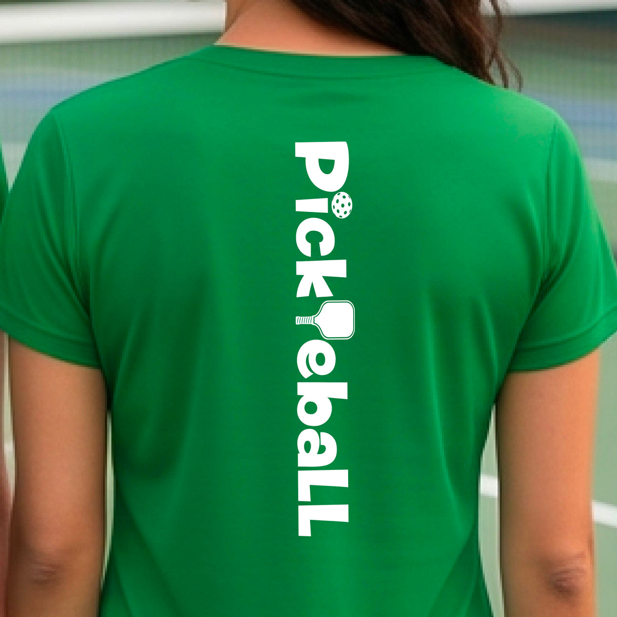 A woman wearing a green short-sleeve t-shirt with a white graphic text 'PICKLEBALL' with a pickleball paddle replacing the L, on the back, standing on a pickleball court.
