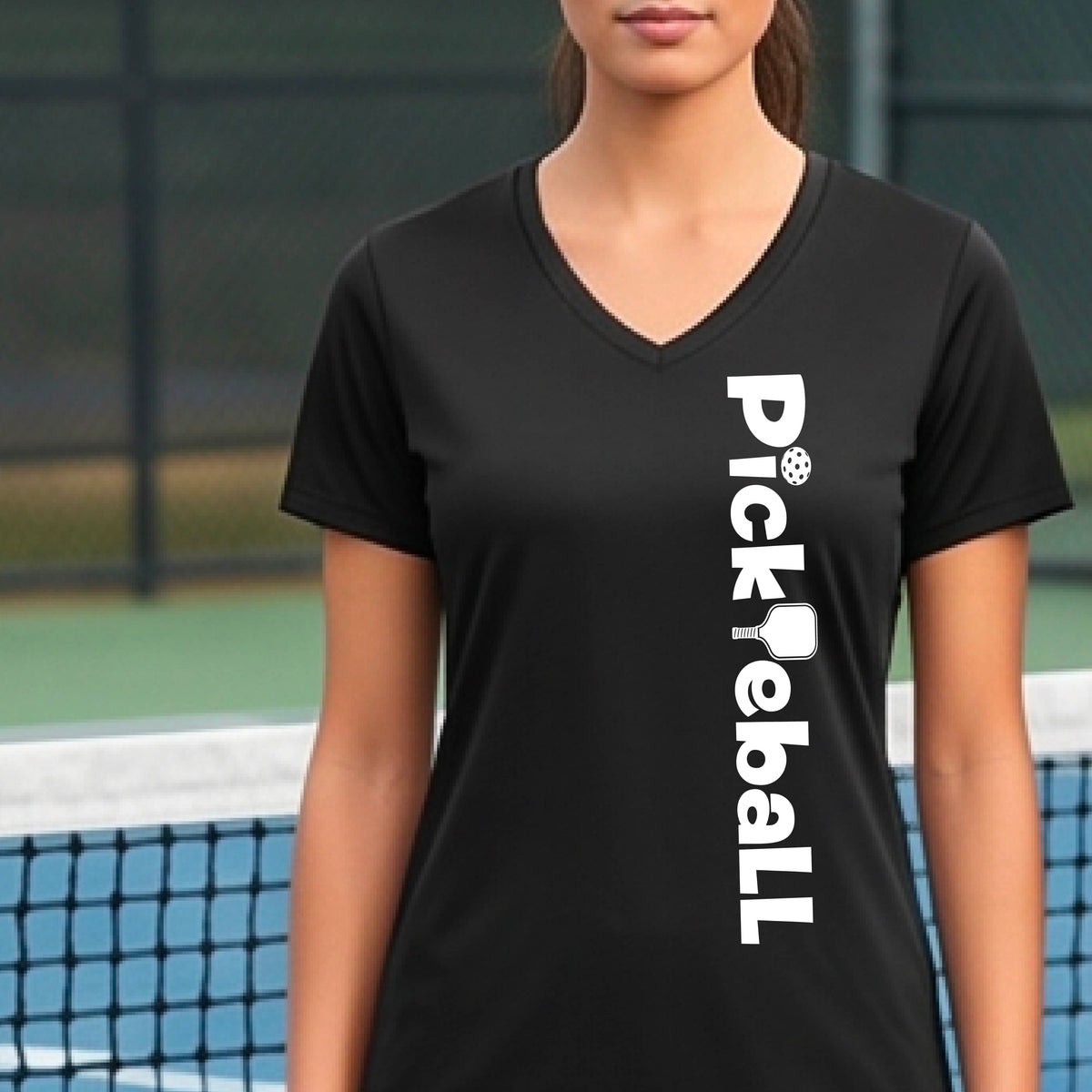 A person wearing a black short-sleeve athletic T-shirt with a vertical white 'Pickbeall' logo printed on the front right, photographed on a blue pickleball.