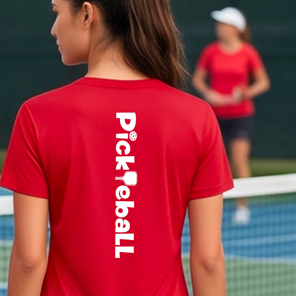 A person wearing a red short-sleeve athletic T-shirt with a vertical white 'Pickbeall' logo printed on the middle back, photographed on a pickleball court.