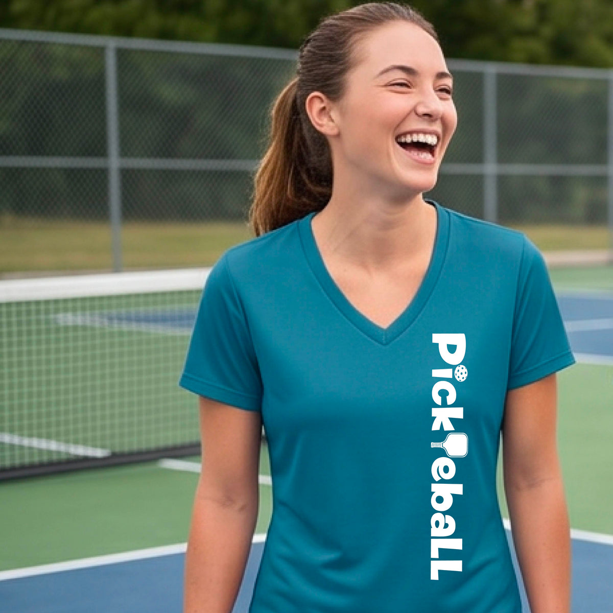 A person wearing a teal short-sleeve athletic T-shirt with a vertical white 'Pickbeall' logo printed on the front right, photographed on a pickleball court.