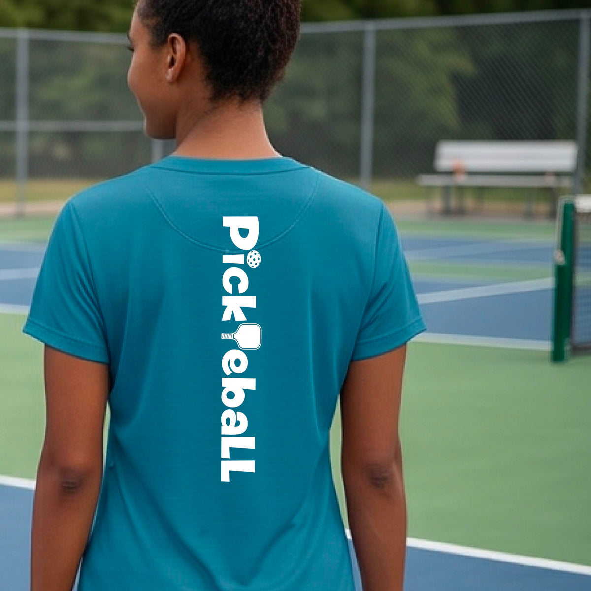 A person wearing a teal short-sleeve athletic T-shirt with a vertical white 'Pickbeall' logo printed on the middle back, photographed on a pickleball court.