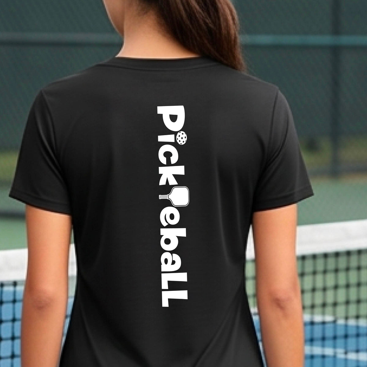 A person wearing a black short-sleeve athletic T-shirt with a vertical white 'Pickbeall' logo printed on the middel back, photographed on a blue pickleball.