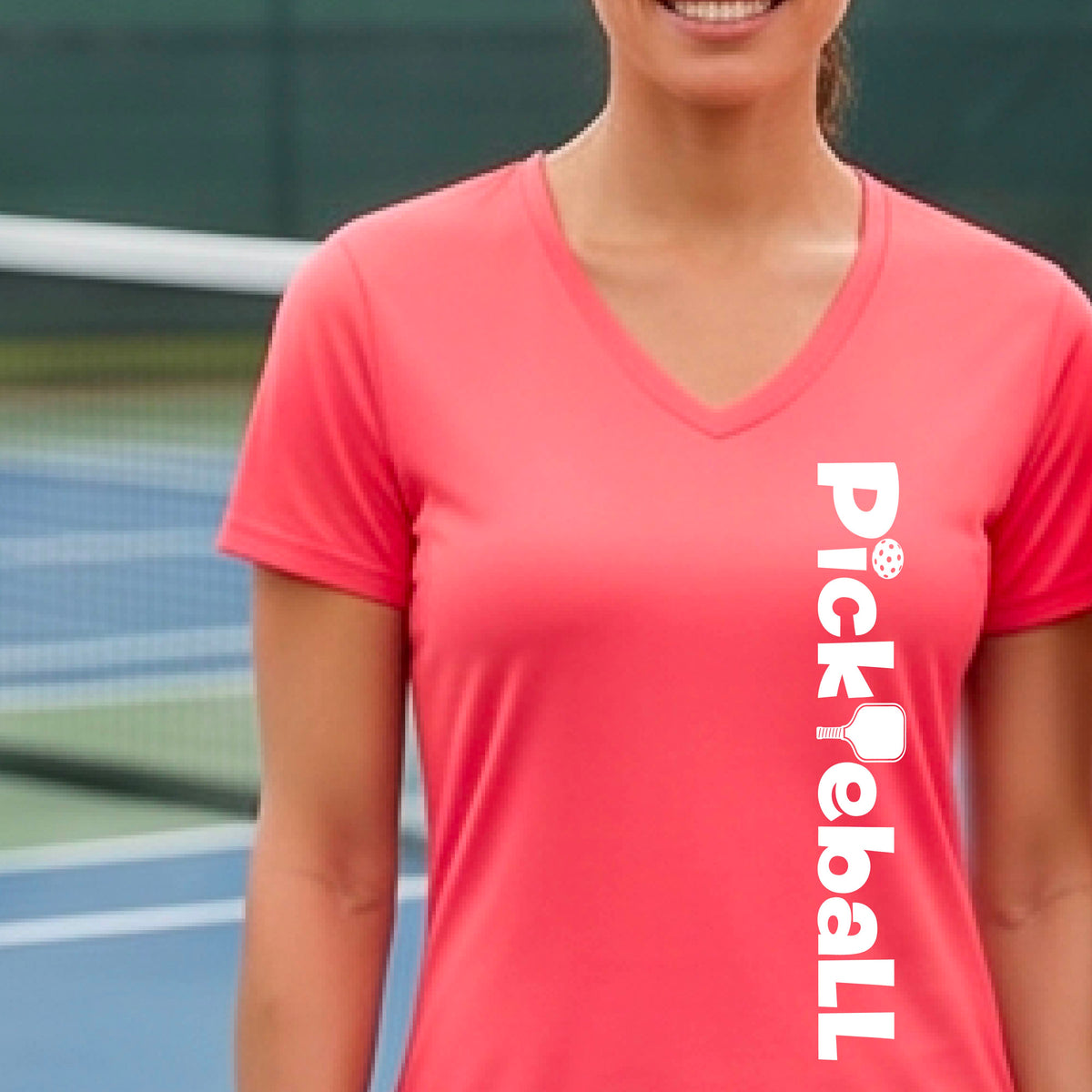 A person wearing a coral pink short-sleeve athletic T-shirt with a vertical white 'Pickbeall' logo printed on the front right, photographed on a pickleball court.