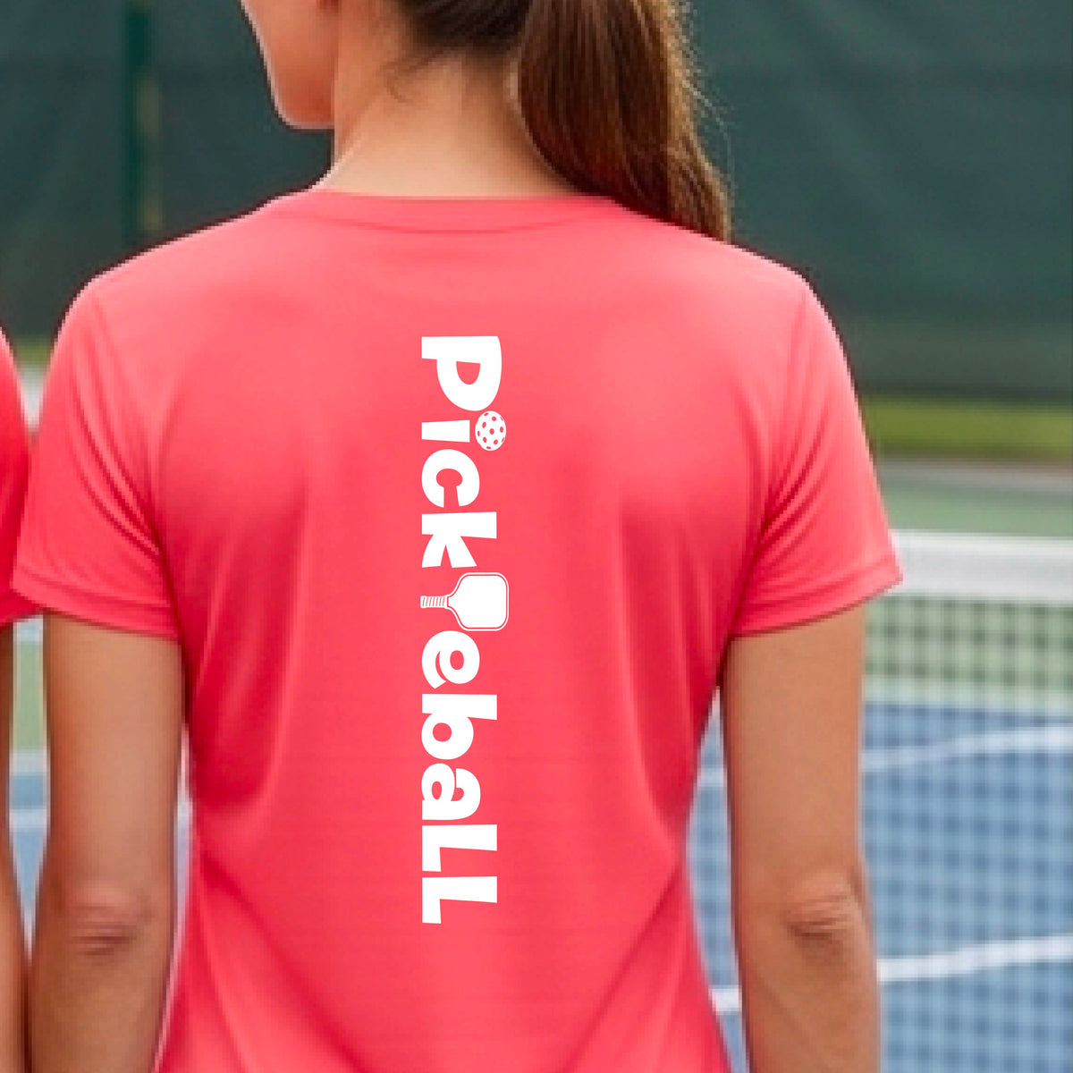 A person wearing a coral pink short-sleeve athletic T-shirt with a vertical white 'Pickbeall' logo printed on the middle back, photographed on a pickleball court.