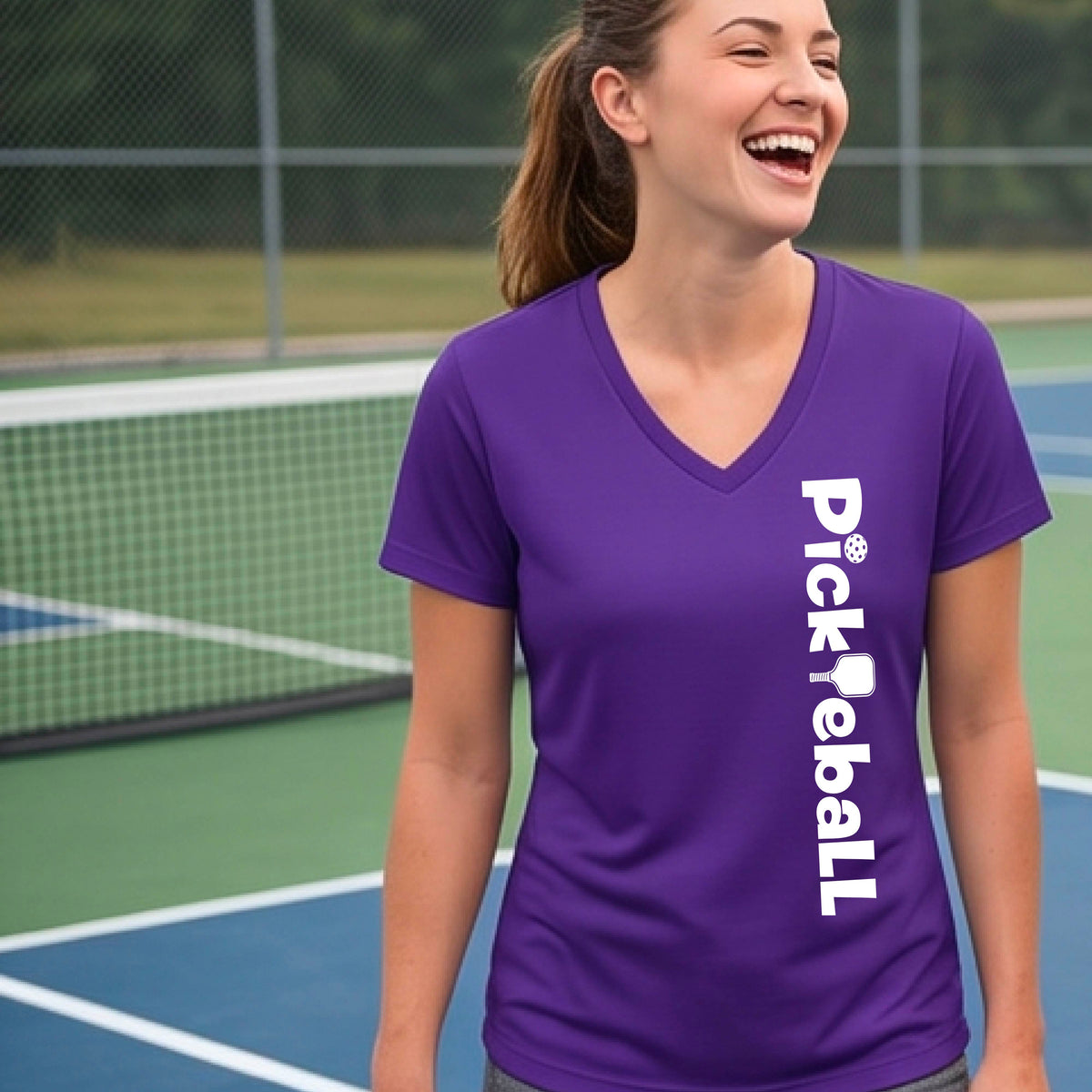 A person wearing a purple short-sleeve athletic T-shirt with a vertical white 'Pickbeall' logo printed on the front right, photographed on a pickleball court.