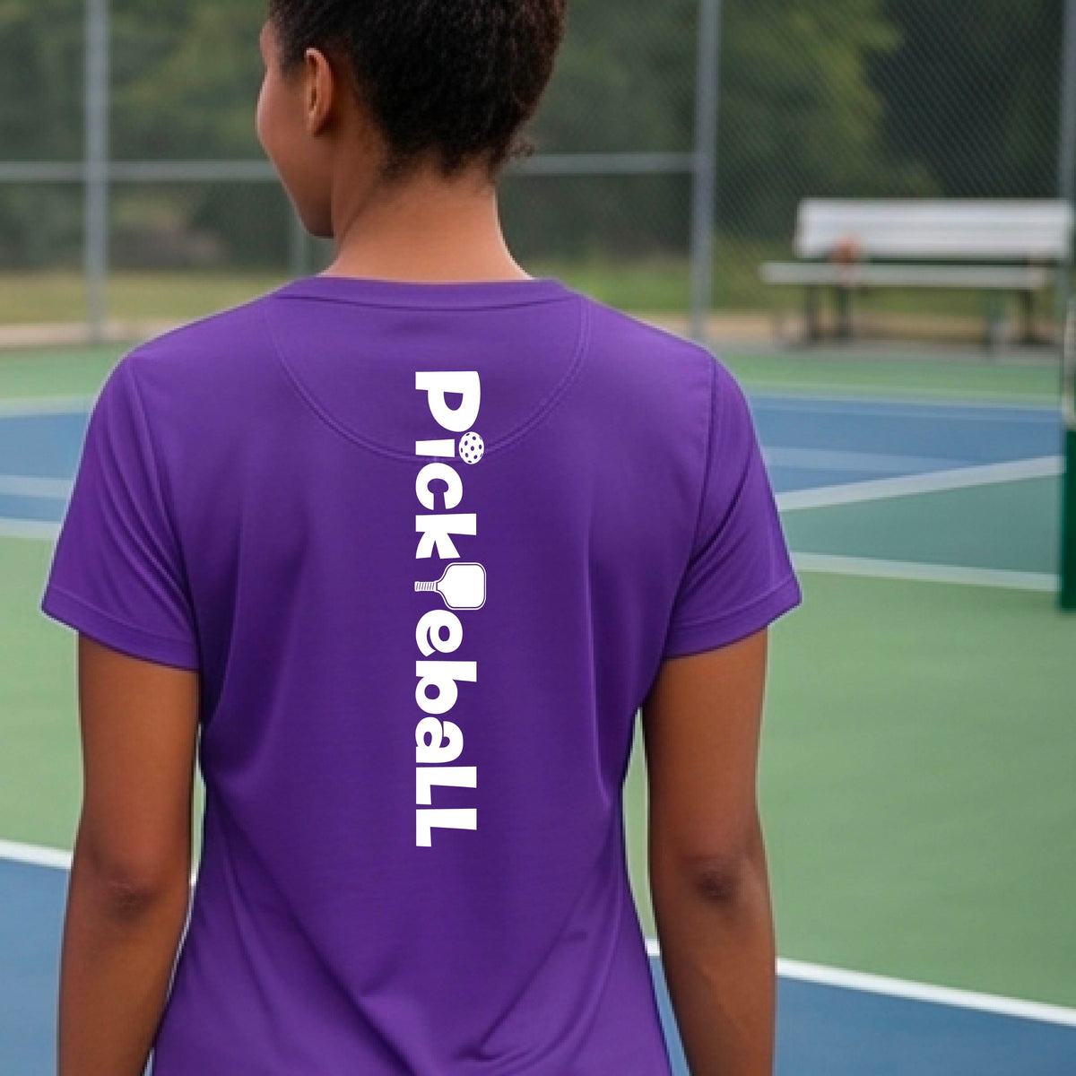 A person wearing a purple short-sleeve athletic T-shirt with a vertical white 'Pickbeall' logo printed on the middle back, photographed on a pickleball court.