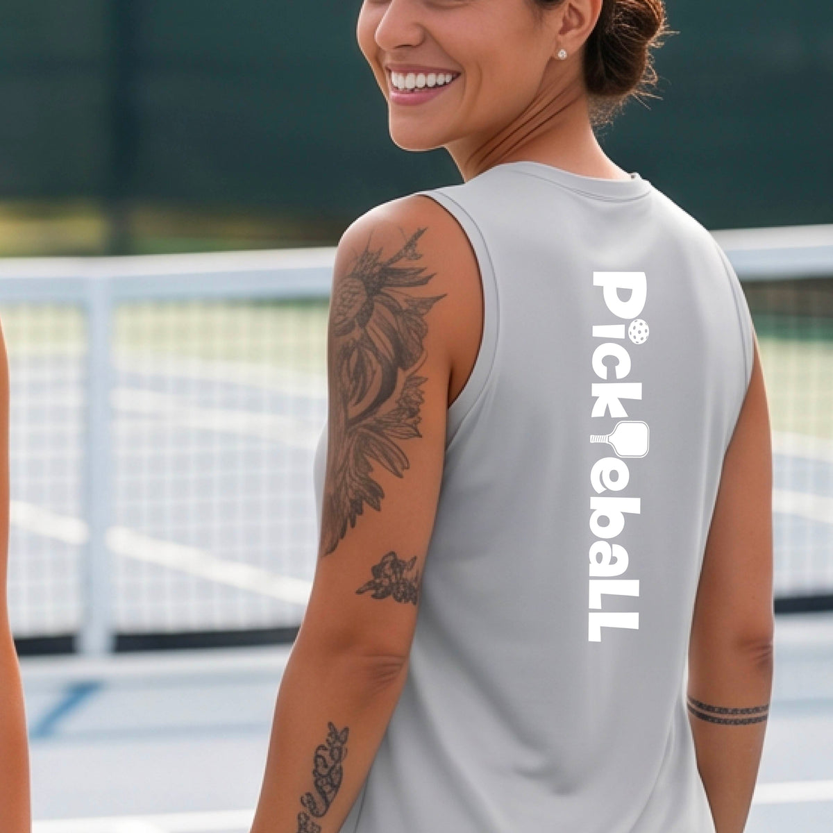 A smiling woman on a pickleball court wearing a bright silver sleeveless V-neck athletic top with 'Pickleball' printed vertically in white on the back.