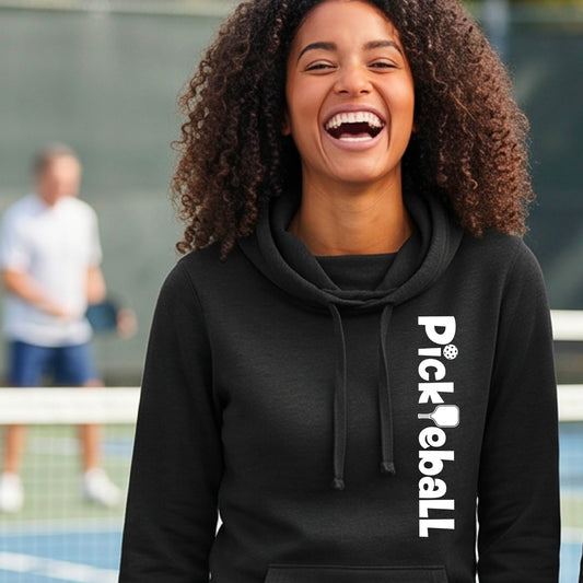 A woman smiling broadly, wearing a black cowl-neck hoodie with 'Pickleball' printed vertically in white on the right side, set against a blurred pickleball court background.