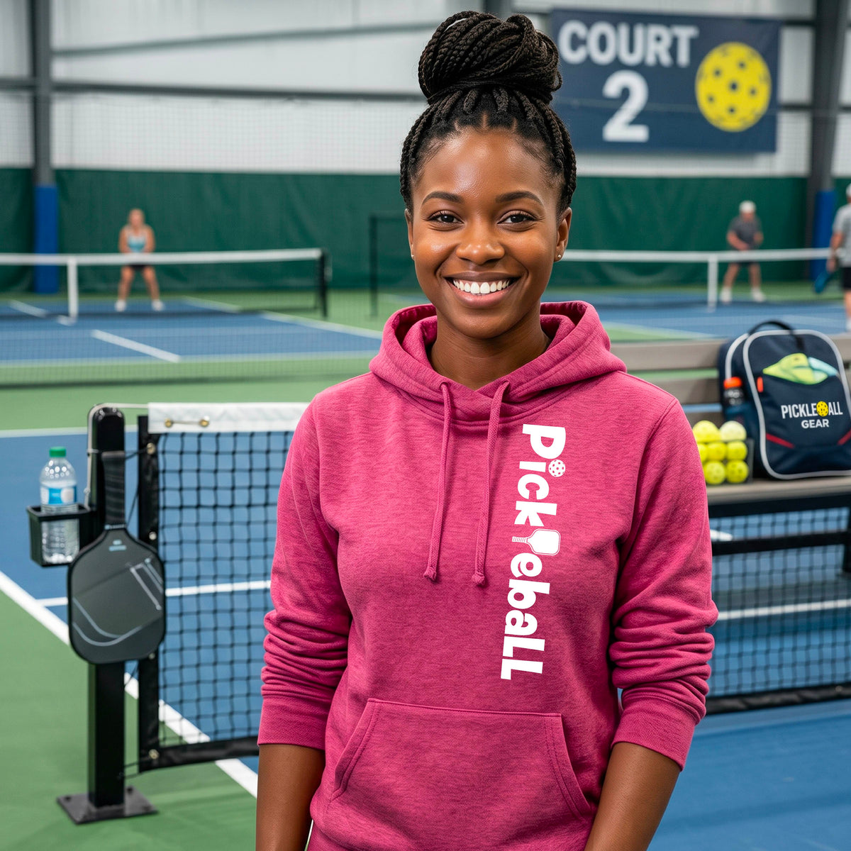 A smiling woman stands on an indoor pickleball court wearing a heathered pink cowl-neck hoodie with 'Pickleball' printed vertically in white on the front. Pickleball paddles, balls, and a branded bag are visible around her, with court nets and other players in the background.