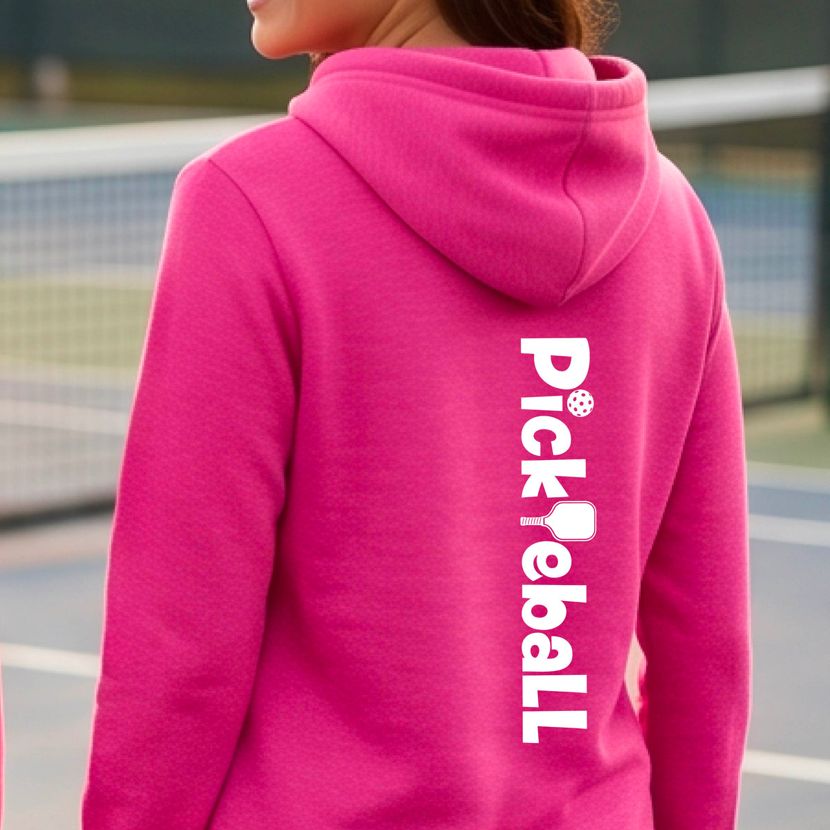A person wearing a bright pink hoodie, shown from the back, with the word 'Pickleball' printed vertically in white on the back. The background is a blurred outdoor pickleball court.