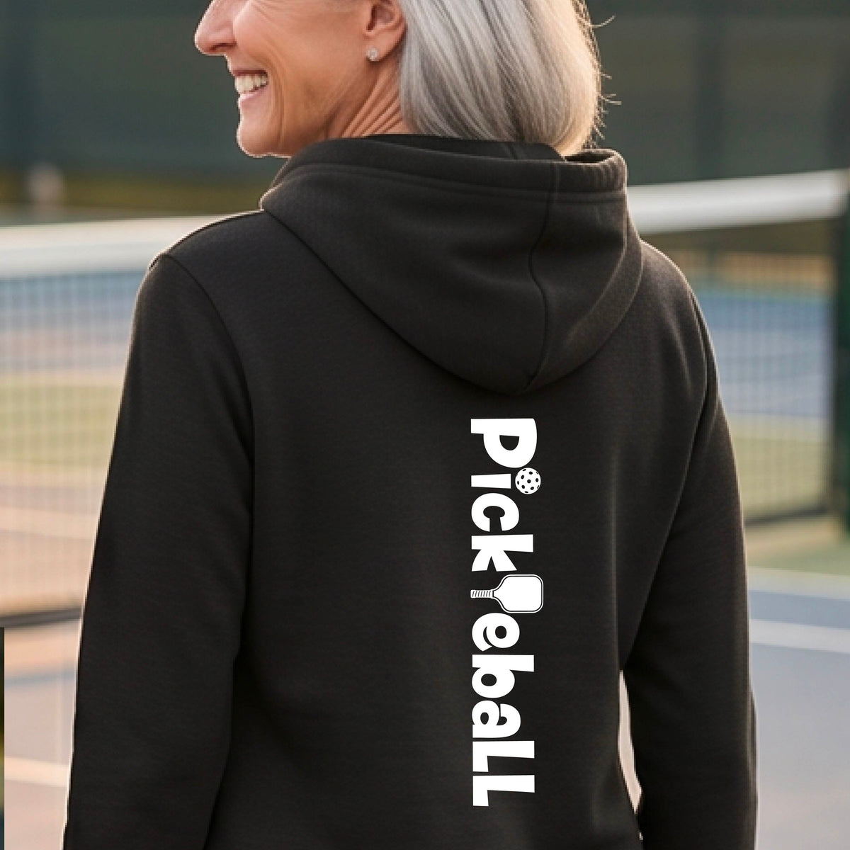 A woman with short gray hair is shown from the back wearing a black hoodie with 'Pickleball' printed vertically in white on the back, standing on a tennis court.