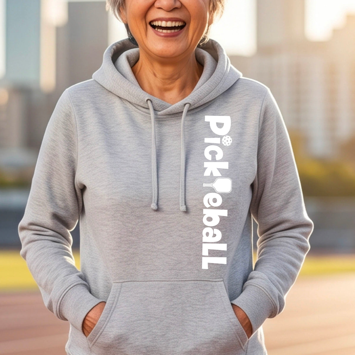 A smiling older woman wearing a light heather gray pullover hoodie with a vertical white 'Pickleball' graphic on the left side, standing outdoors with a blurred city background.