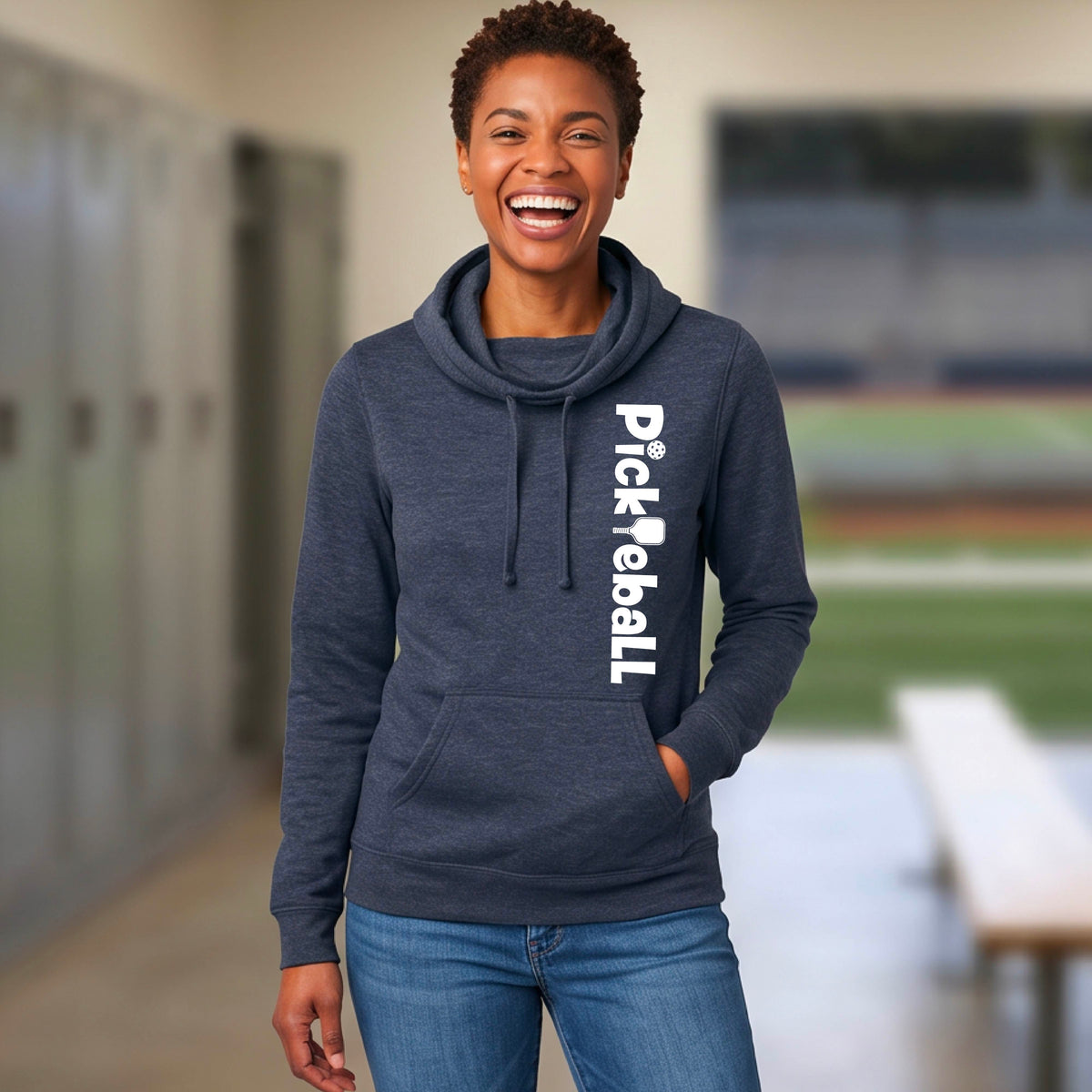 A woman smiling in a school hallway wearing a dark heather gray hoodie with 'Pick 'PickleBall' printed vertically in white on the right side, paired with blue jeans.