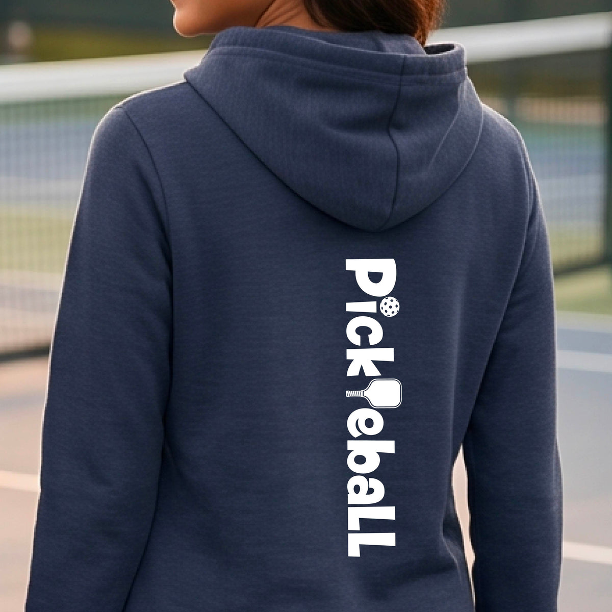 A person viewed from the back wearing a navy blue hoodie with 'Pickleball' printed vertically in white on the back, standing on an outdoor pickleball court.
