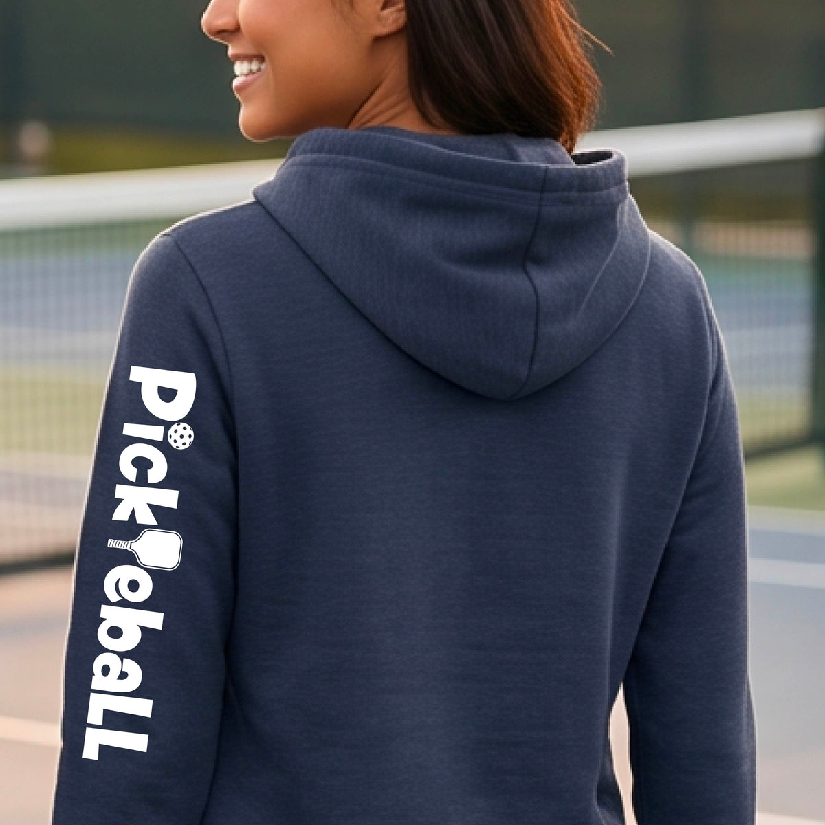 A woman wearing a navy blue hoodie with 'Pickleball' printed in white down the left sleeve, standing on an outdoor pickleball court.