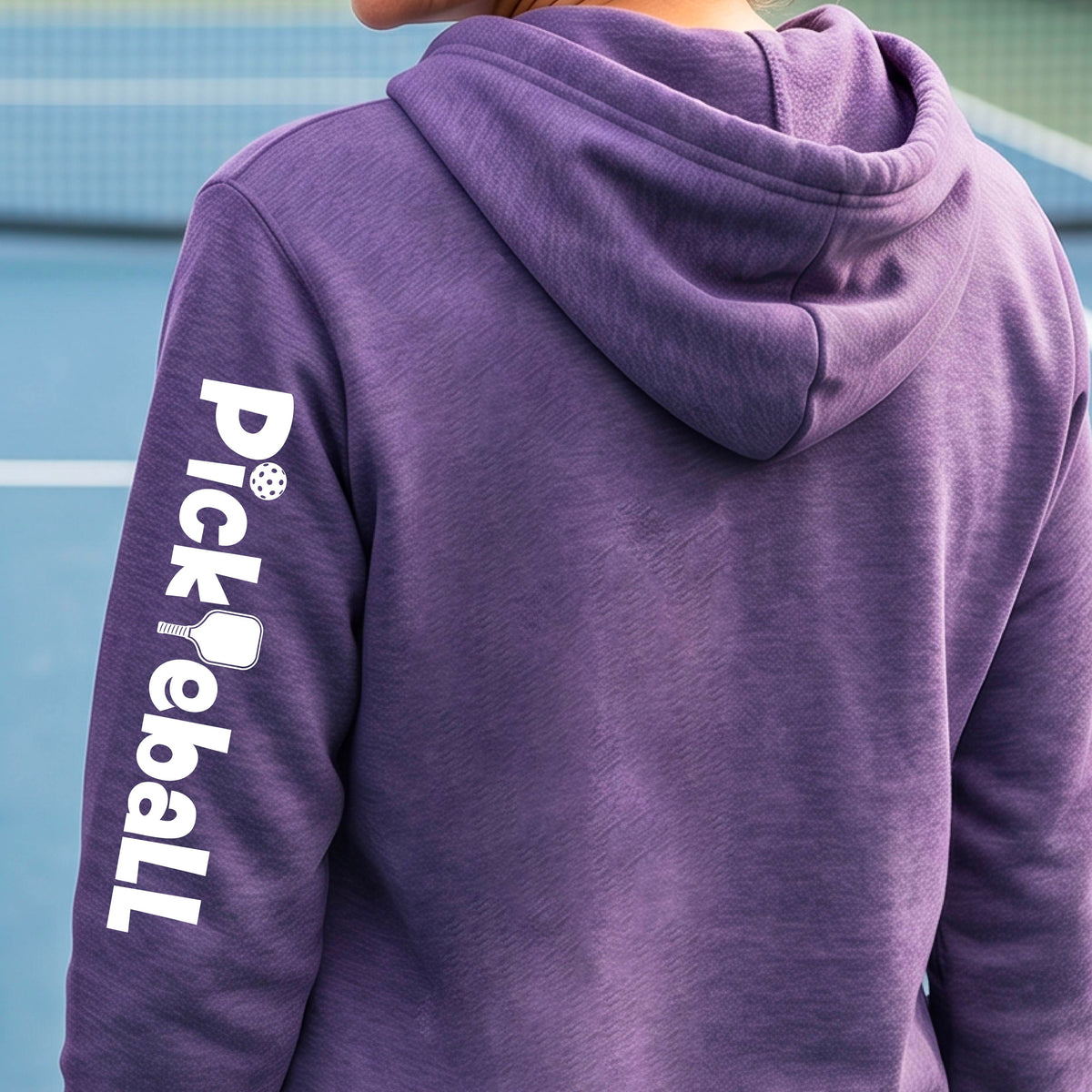 A person wearing a heather purple hoodie from the back, standing on a blue tennis court. The left sleeve features a vertical white graphic that reads 'Pickball' with a pickleball icon.