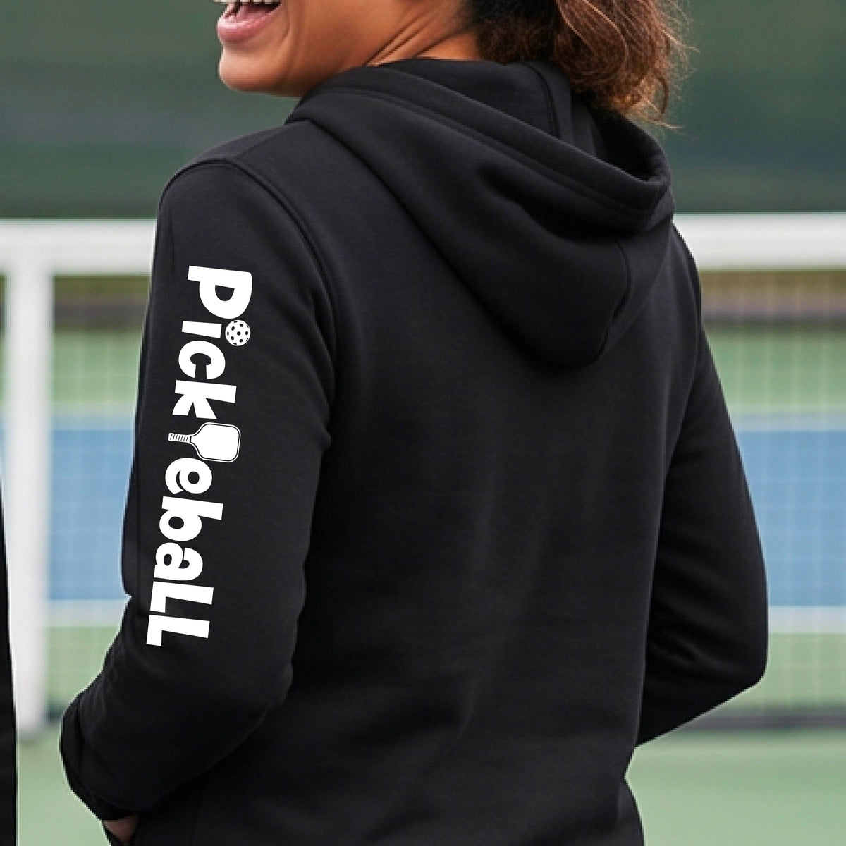 A person wearing a black hoodie with 'Pickleball' printed vertically in white on the left sleeve, photographed outdoors on a sports court.