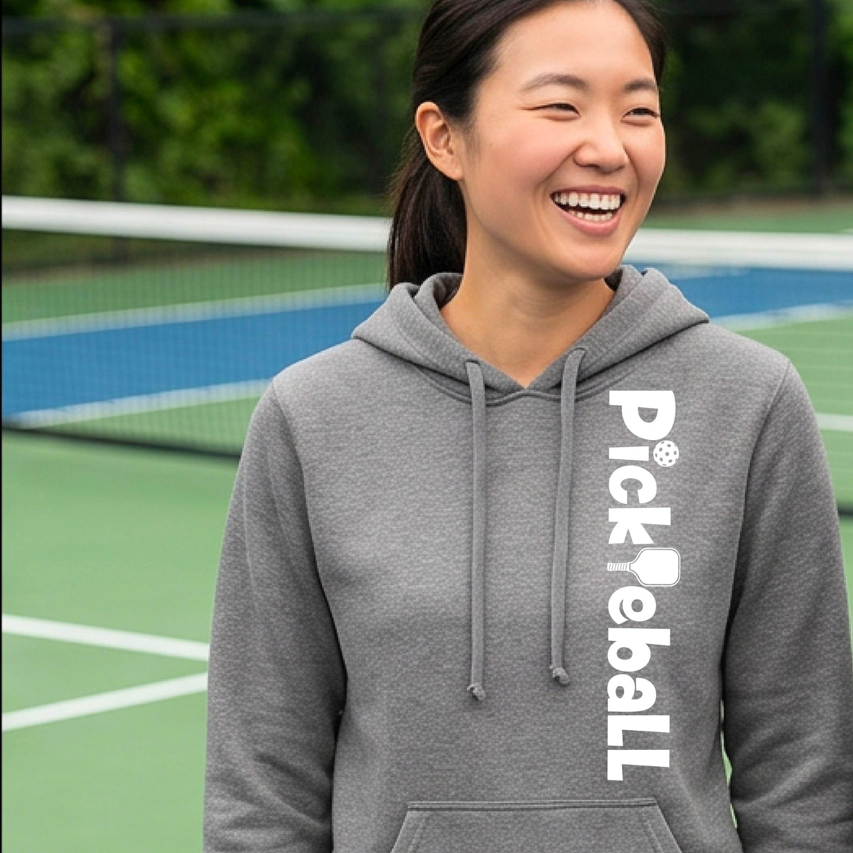 A woman smiling on a pickleball court wearing a heather gray hoodie with 'Pickleball' printed vertically in white on the front.