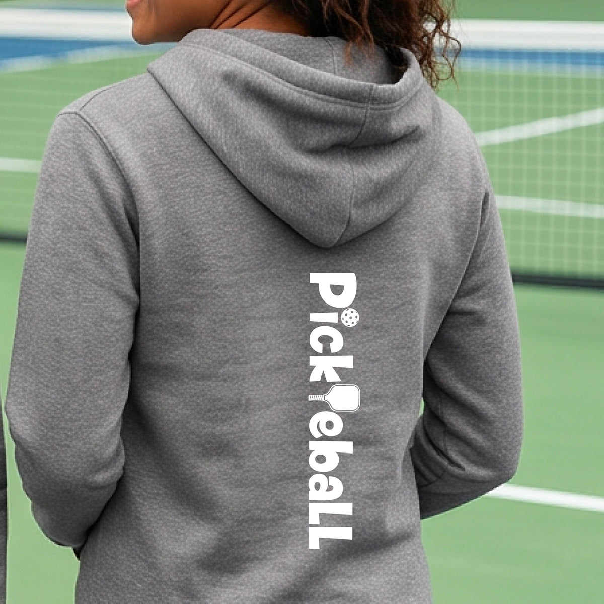 A person viewed from behind wearing a heather gray hoodie with 'Pickleball' printed vertically in white on the back, standing on a green pickelball court.