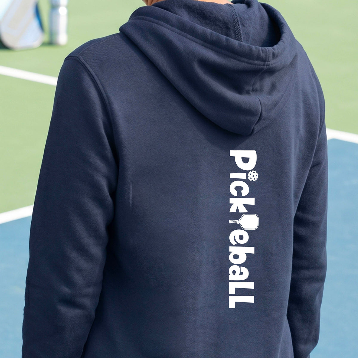 A person wearing a navy blue hoodie on a pickelball court, shown from the back. The hoodie features the word 'Pickleball' printed vertically in white on the back, with a pickleball and paddle graphic replacing the 'i' in the word.