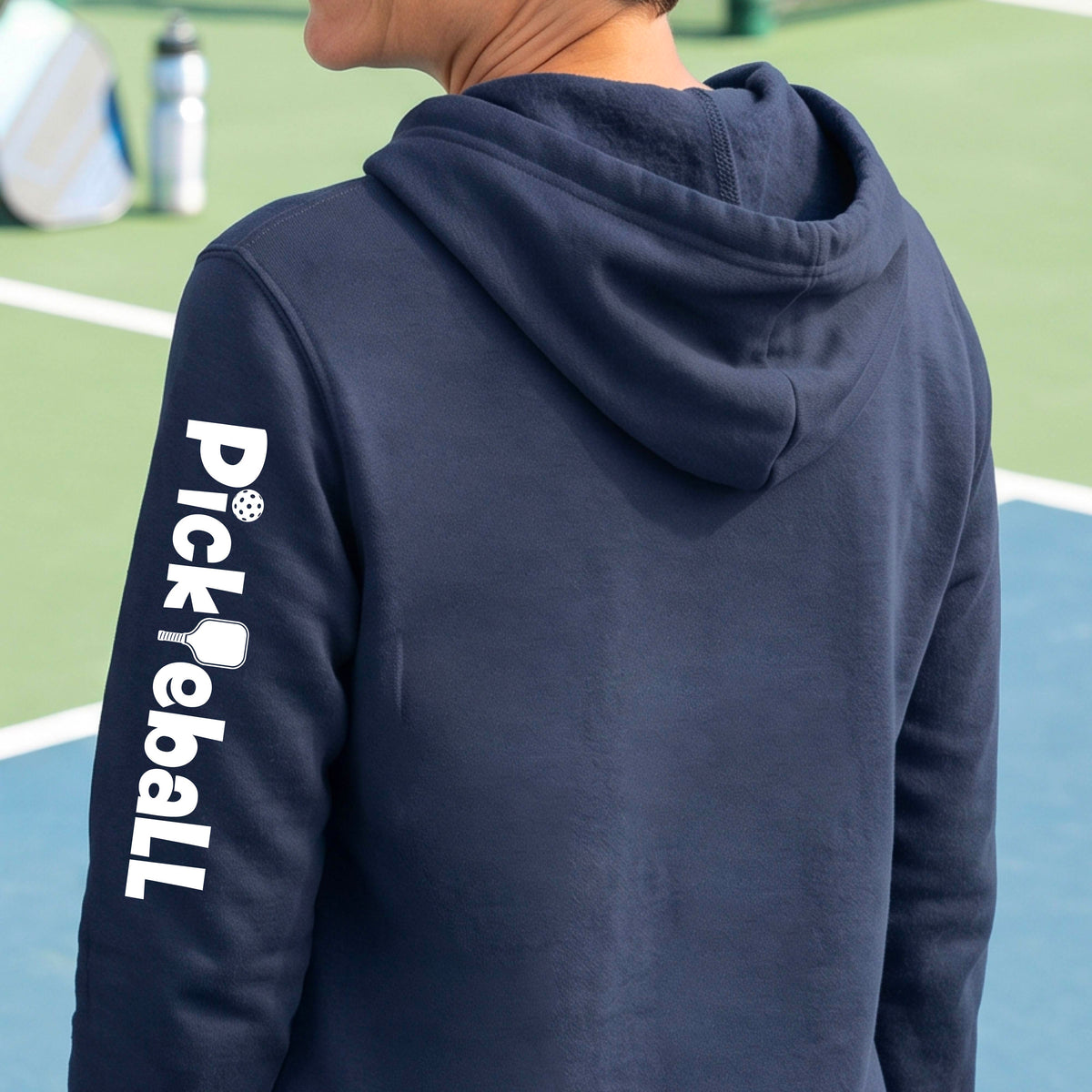 A woman wearing a navy bluepullover hoodie with 'Pickleball' printed vertically in white on the sleleve, standing on a blue pickleball court.