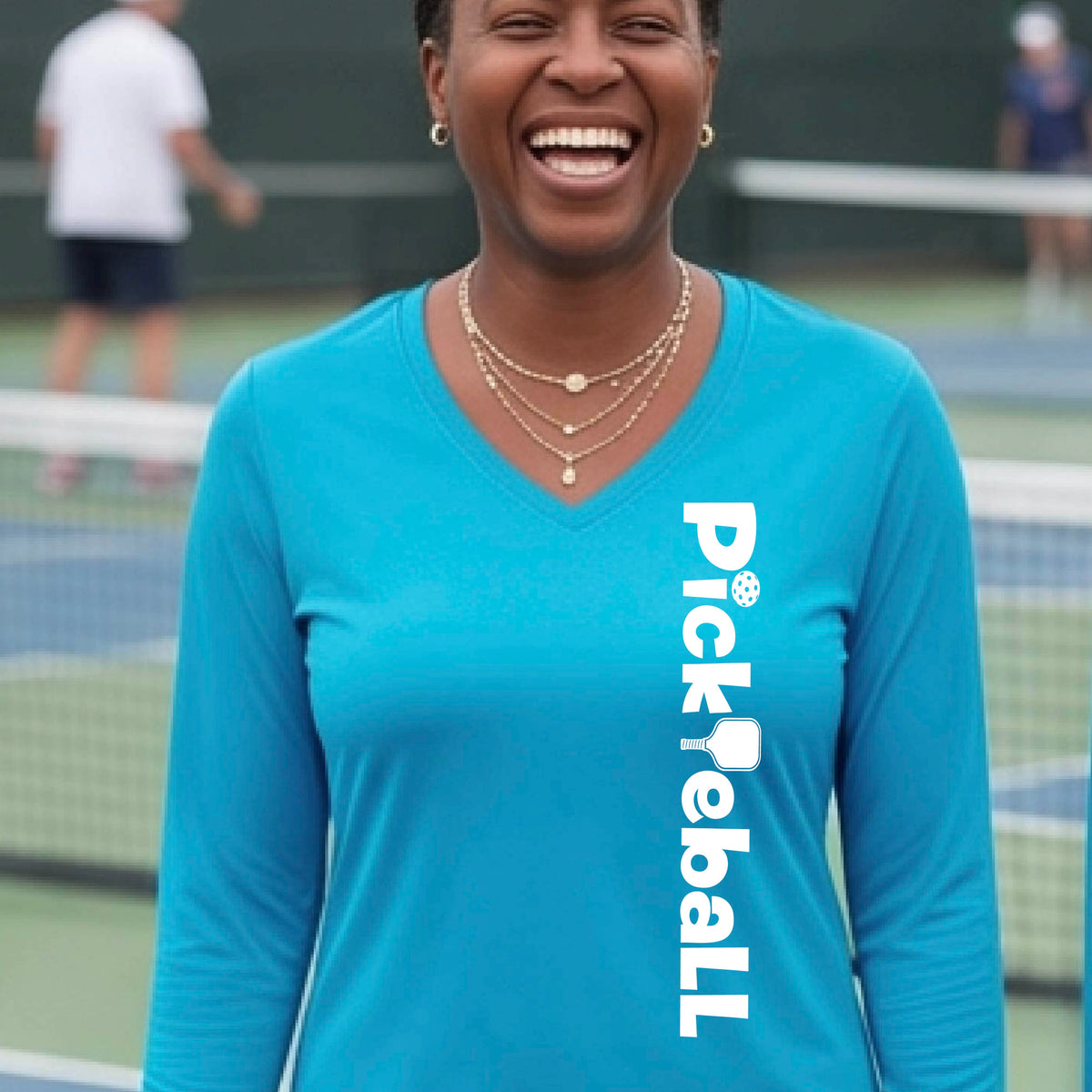 A woman wearing a bright blue long-sleeve t-shirt with a white graphic text 'PICKLEBALL' with a pickleball paddle replacing the L, on the front, standing on a pickleball court.