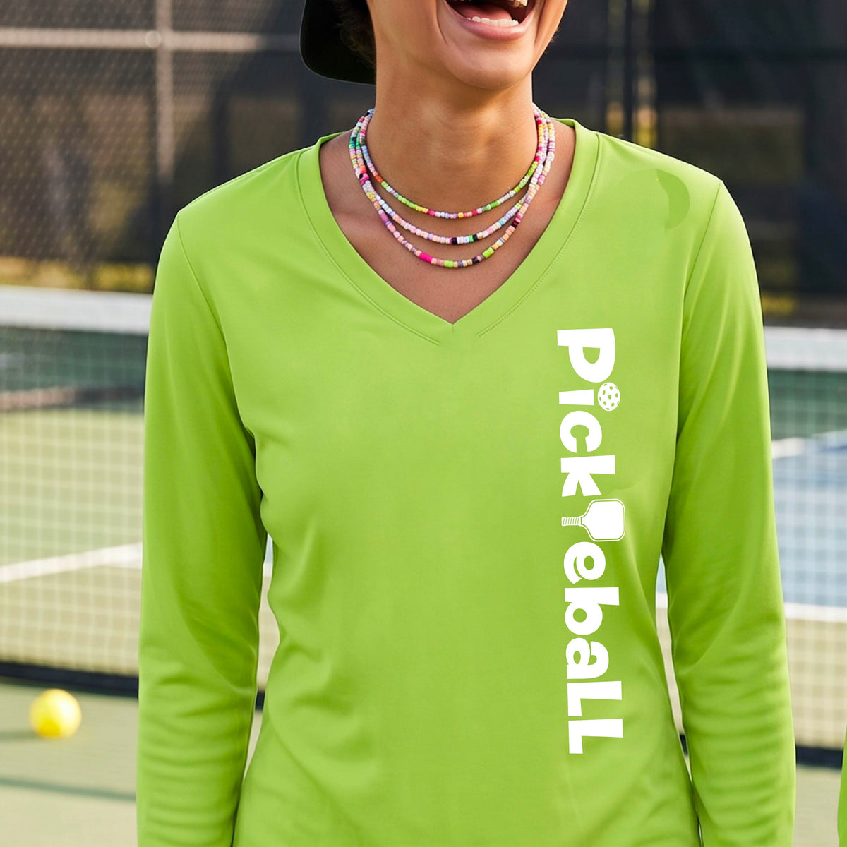 A woman wearing a lime green long-sleeve t-shirt with a white graphic text 'PICKLEBALL' with a pickleball paddle replacing the L, on the front, standing on a pickleball court.