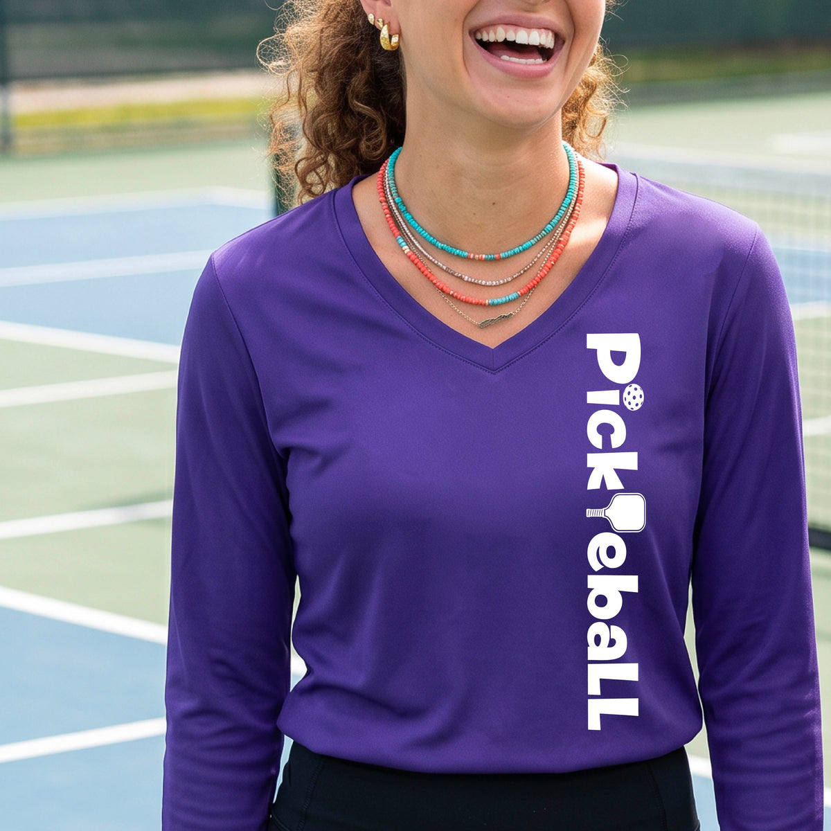 A woman wearing a purple long-sleeve t-shirt with a white graphic text 'PICKLEBALL' with a pickleball paddle replacing the L, on the front, standing on a pickleball court.